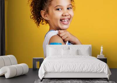 Happy, portrait and child with a plaster in a studio for a wound, sore or injury on her arm. Happiness, smile and healthy girl kid model posing with a bandaid after a vaccination by yellow background Wall mural