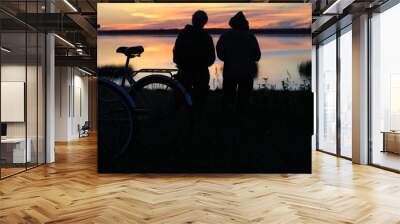 Two bicycles on the high shore of the lake in front of people looking ahead at the sunset at the end of the day Wall mural