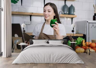 Young beautiful woman using laptop computer searching and learning for cooking healthy food from fresh vegetables and fruits in kitchen room. Wall mural