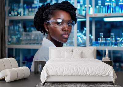 A female scientist in a lab coat and safety goggles analyzes data on a computer screen. Behind her, shelves filled with beakers and other lab equipment line the walls Wall mural