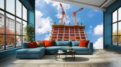 Two construction cranes rise against a blue sky with white clouds, working on a tall, unfinished skyscraper. Wall mural