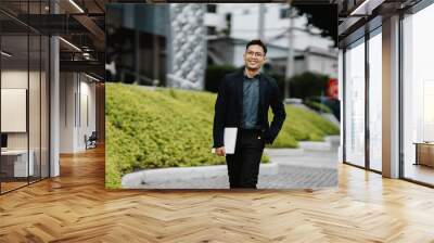 Confident Businessman: A portrait of a smiling Asian businessman striding confidently outdoors, holding a tablet. He exudes success and professionalism. Wall mural