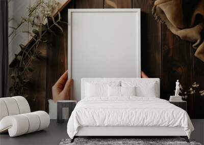 Girl holds a white poster with white frame mockup in her hands with green plants around on wooden background Wall mural