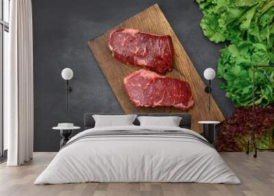 two raw pieces of beef steak on a brown wooden board, black table, next to green lettuce leaves Wall mural