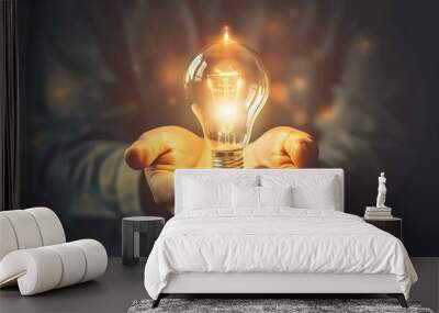 A glowing lightbulb surrounded by two hands gently holding it, with an upward arrow emerging from the bulb, symbolizing the growth of ideas and innovation on a dark, moody background Wall mural