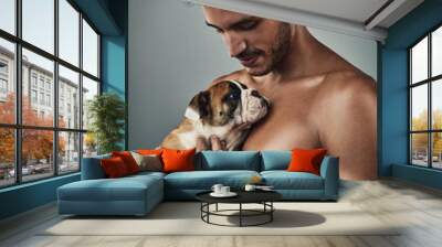 Cuteness just upped its game. Studio shot of a handsome and shirtless young man holding a bulldog puppy against a grey background. Wall mural