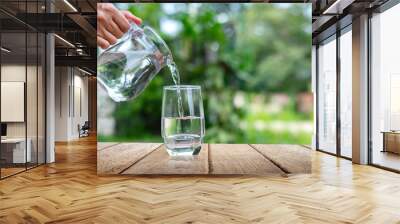 Water from jug pouring into glass on wooden table outdoors.Drink water pouring in to glass over sunlight and natural green background.Photo select focus with copy space. Wall mural