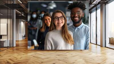 Two happy entrepreneurs smiling at the camera cheerfully. Young businesspeople standing in a boardroom with their colleagues in the background. Diverse entrepreneurs working together as a team. Wall mural