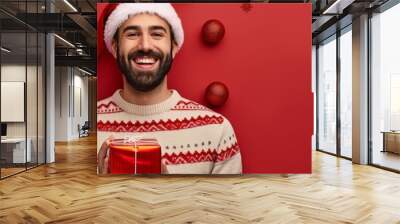 A cheerful man in a festive Santa hat holds a wrapped gift against a vibrant red backdrop with Christmas ornaments hanging around. Wall mural