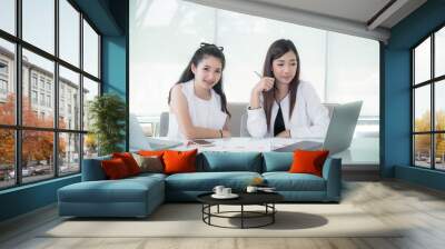 Young happy friends working in office. Portrait of two beautiful Asian woman sitting in office with laptop and document paper chart on desk. Wall mural