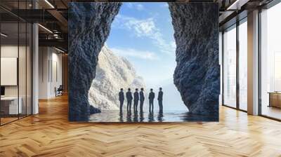 Silhouetted figures stand inside a large rock arch, overlooking a scenic view of the ocean and a clear blue sky. Wall mural