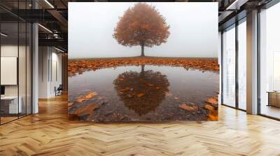 solitary tree with vibrant orange leaves stands in foggy landscape, reflected in puddle surrounded by fallen leaves, creating serene autumn scene Wall mural