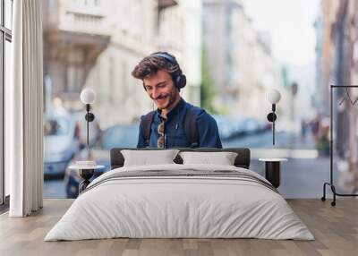 Positive young man holding phone and listening music on headphones at street Wall mural