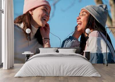 two girls looking happy while sharing a smartphone and earphones, outside and wearing casual clothes Wall mural