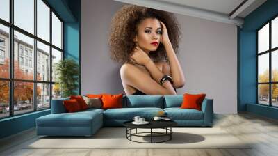 Beautiful young African American woman with afro in a fresh dark short summer dress posing holding up one edge of the flared skirt with a provocative expression, on a dark grey background Wall mural