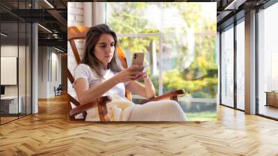 Woman using the smartphone sitting in a rocking chair on the window with garden in the background Wall mural