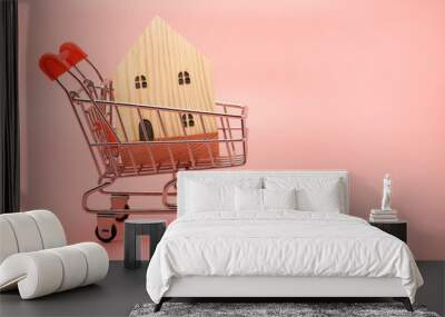 Miniature decorative wooden house in a shopping cart trolley isolated on pastel pink background with copy space. Sale and purchase of real estate concept. Wall mural