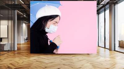 Coughing Asian woman wearing mask and holding blue umbrella on pink background. Wall mural