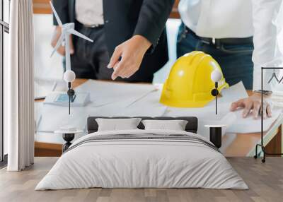 A meeting of two male engineers in an office is pointing at a wind turbine model regarding the future design of the energy industry. On the table are hard hats, blueprints, pens. Wall mural