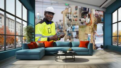 At the industrial plant, American male engineers in helmets and vests meticulously analyze circuit boards through close-up inspections, utilizing tablets for accurate diagnostics. Wall mural