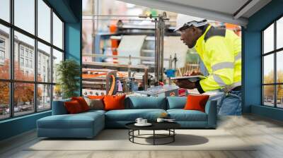 At the industrial plant, the diligent American male engineer, in a helmet and vest, employs a tablet to troubleshoot and optimize the machine, ensuring smooth production. Wall mural