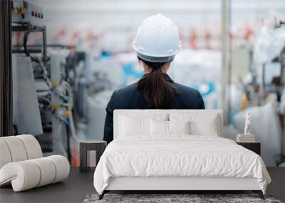 back photo of an Asian female engineer standing in the middle of a large plastic and steel industrial factory wearing a hard hat and suit. Around there are machines working in full system. Wall mural