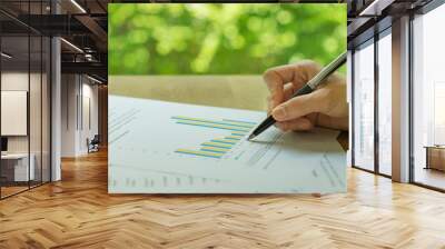 Business, investment or financial report review, hand holding pen reviewing bar graph information data print on paper with green bokeh in the background Wall mural