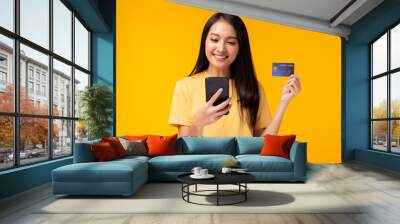 Portrait happy young asian girl standing over yellow background, holding mobile phone, showing credit card. Online payment, hands holding credit card, using smart phone for online shopping	 Wall mural