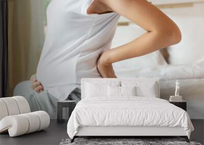 Pregnant woman touching tummy and suffering from back ache sitting on bed at home. Young attractive pregnant mother get back pain, her fetus or baby and belly bigger than before that mom get painful Wall mural