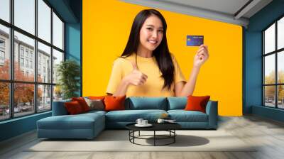 Young asian woman showing plastic credit card giving thumb up to credit card, standing over yellow background. Beautiful customer girl get satisfied of credit card service and promotion, smile face Wall mural