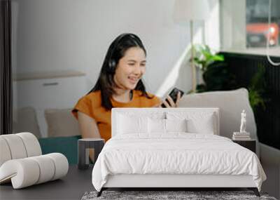 Smiling girl relaxing at home, she is playing music using smartphone tablet, laptop, and wearing white headphones. . Wall mural