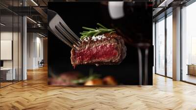 Close-up of a rib-eye steak on a fork, garnished with rosemary and sea salt, with a glass of red wine. The black background creates a luxurious dining scene. Wall mural