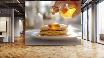 Close-up of a woman's hand pouring honey over golden pancakes on a white plate, set on a white table with a softly blurred background, capturing a serene and appetizing breakfast moment Wall mural