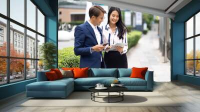 Asian business man talking on mobile phone and young beautiful woman working on digital tablet computer standing outside office building. Wall mural