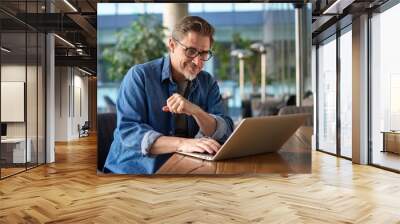 Happy middle age businessman using laptop computer in office. Older casual man working online on terrace of cafe, smiling. Wall mural