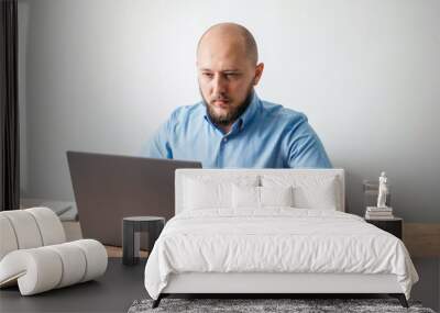 Bald young man with beard working online on his laptop at the wooden table in a home office with white wall as background. Business, freelance or study concept Wall mural