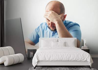 Frustrated and tired young man working online on his laptop at the wooden table in a home office, feels headache, stress and problems. Business, freelance or study concept Wall mural