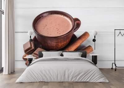 Hot cocoa with milk in brown clay cup, broken chocolate cubes, hazelnut and cinnamon sticks on white wooden planks Wall mural