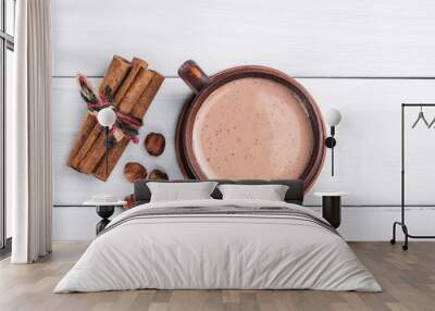 Hot cocoa with milk in brown clay cup, hazelnut and cinnamon sticks on table of white wooden planks, top view Wall mural