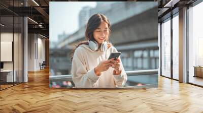 Beautiful young happy asian woman with headphones looking at mobile phone while standing in the city Wall mural