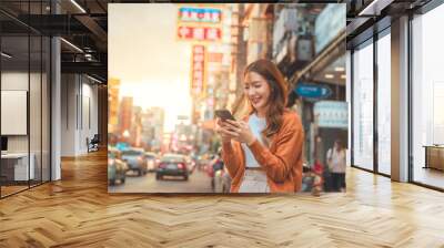 Happy young Asian tourist woman using smartphone on street, Female traveller in Yaowarat Chinatown street food market during sunset in Bangkok, Thailand. Wall mural