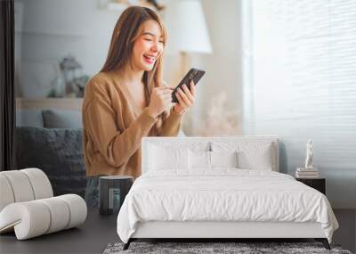 Happy young asian woman browsing surfing wireless internet on mobile phone while sitting a couch in living room at home, Shopping online via website Wall mural