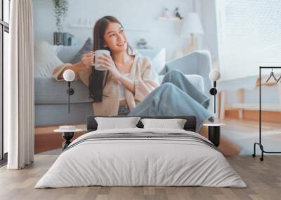 Happy young asian woman drinking coffee relaxing on the rug beside to the sofa at home. Smiling female enjoying resting sitting on couch in modern living room. Wall mural