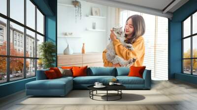 Happy young asian woman hugging cute grey persian cat on couch in living room at home, Adorable domestic pet concept. Wall mural