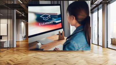 Online education, e-learning. Young woman is sitting at table, working on computer monitor with inscription on screen e-learning and image of square academic cap, distance training. Wall mural