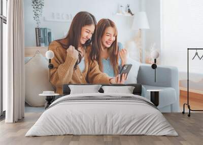 Two excited young asian woman hold phone reading good news feel happy sit on sofa at home, Surprised friends celebrating victory on phone Wall mural