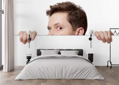 Young man with surprised eyes peeking out from behind billboard paper poster. Man peeking out from the edge and looking at camera isolated on a white background Wall mural