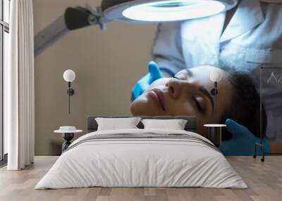 Procedure of facial skin examination at cosmetologist's. Portrait of a young woman with closed eyes and specialist's hands in medical gloves under magnifying lamp. Close up. Indoor shot. Wall mural