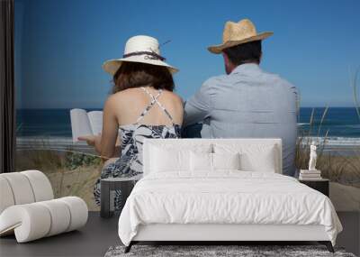 Young girl reading a book at the beach with man wearing a hat Wall mural