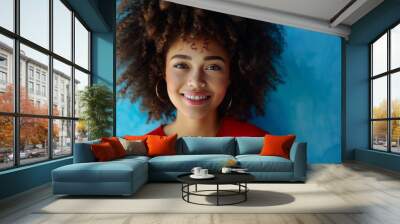 A cheerful woman with curly hair and a red top smiles radiantly in front of a bright blue backdrop, showcasing confidence and joy with natural lighting. Wall mural